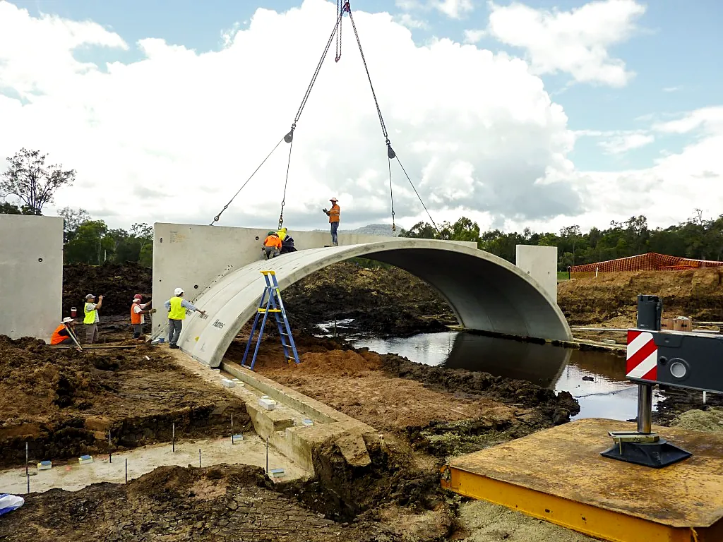 Crew guiding a precast concrete culvert arch into place as a crane lowers it over a creek