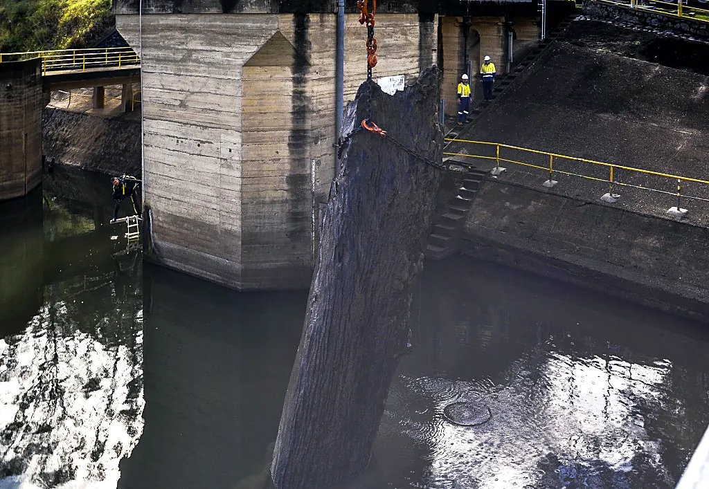 Crane hook lifting a large log out of the water beside a concrete dam wall with a diver and crew