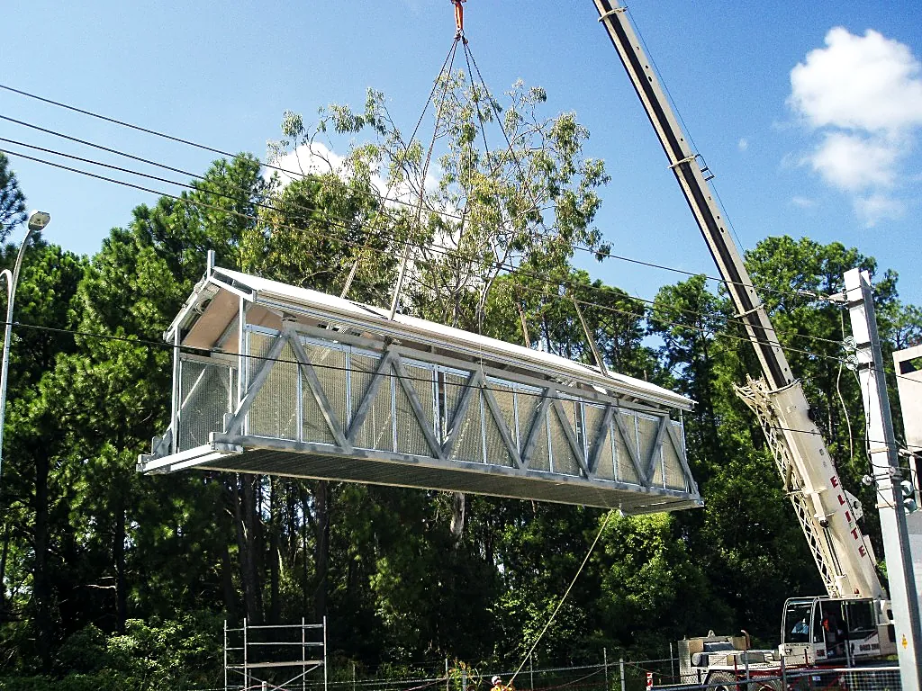 Mobile crane lifting an enclosed steel pedestrian bridge span into position in front of tall trees