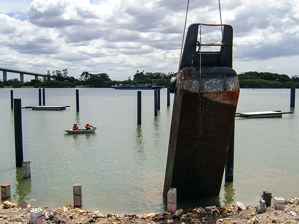 Crane-suspended steel pile guide over a river with piles, pontoons and two workers in a small boat