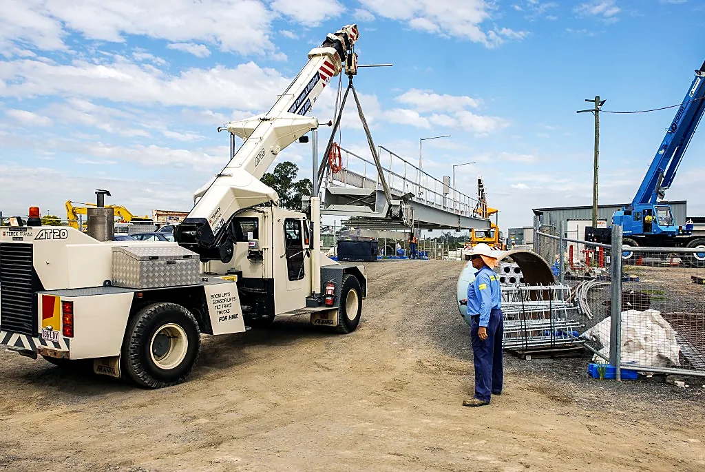 Pick-and-carry crane carrying a steel walkway section across a site while a crew member watches