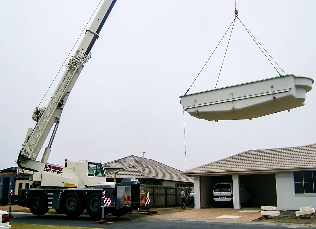 Mobile crane lifting a fibreglass pool shell over a house and garage