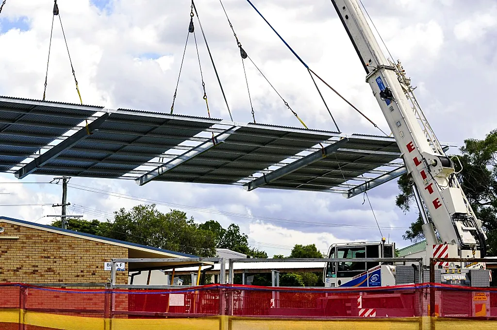 Crane lifting a prefabricated steel and sheeted roof structure beside a brick building